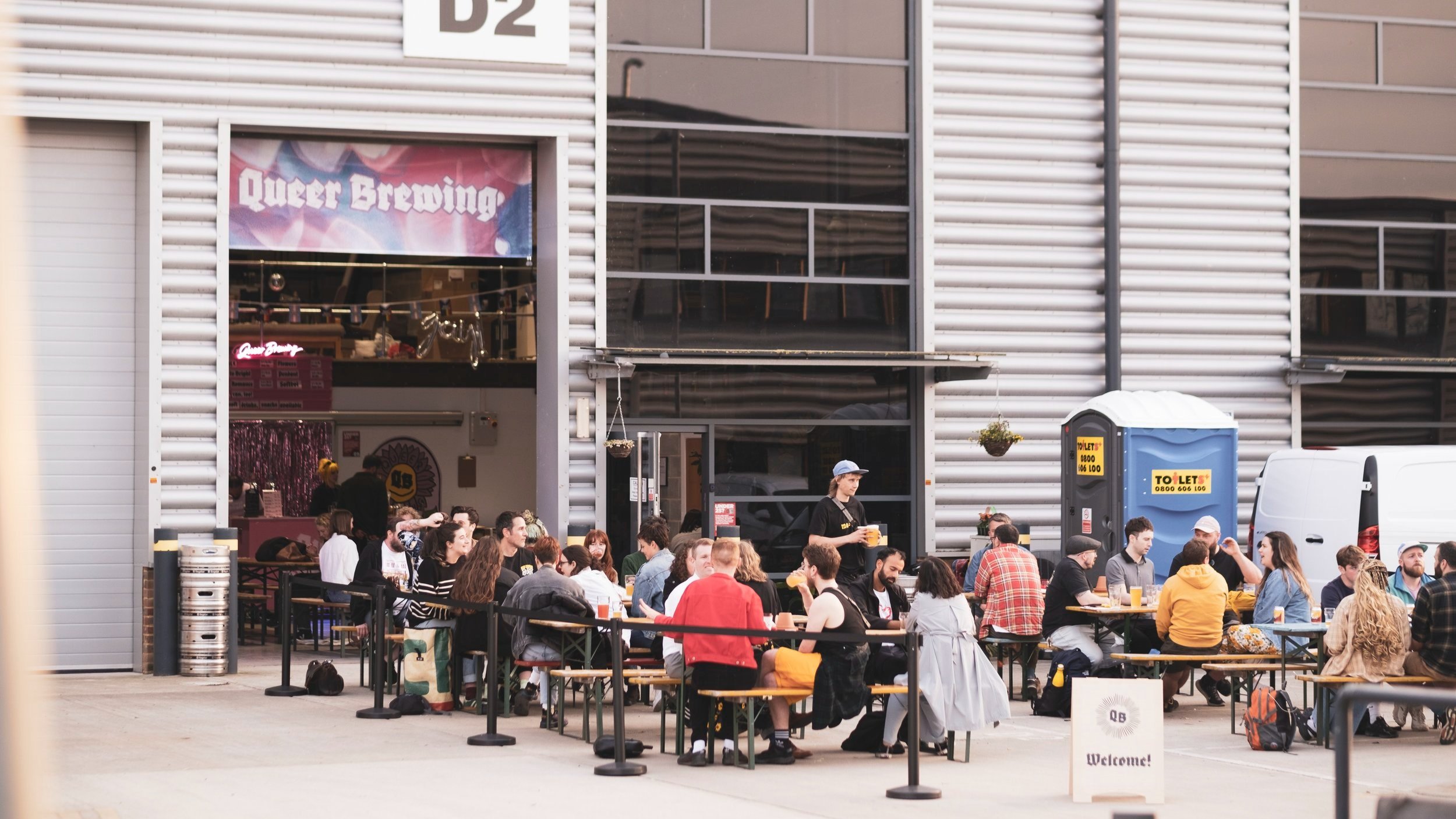 People gathered at outdoor tables, enjoying drinks and conversation in front of an industrial-style building.