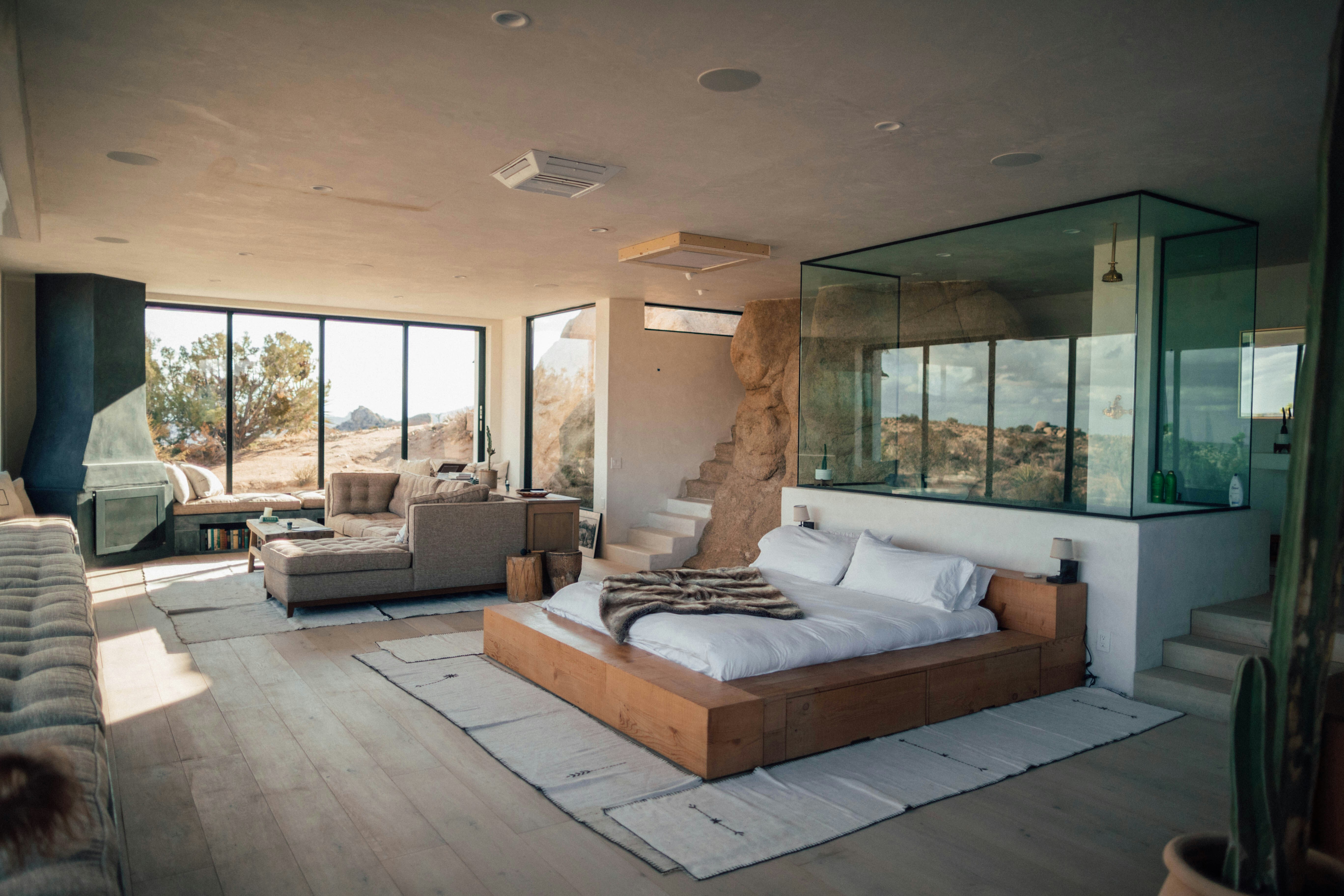 Sunlit interior of a modern desert retreat with minimalist decor, glass-walled shower, and panoramic views of arid landscape through floor-to-ceiling windows.